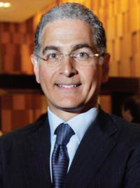 Smiling man with gray hair and glasses in suit and tie against warm blurred background.