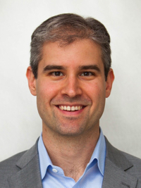 Smiling man with short brown hair wearing a blue shirt and gray blazer on a plain background.