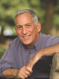 Smiling older man with gray hair leaning on a bench outdoors with green blurred background.
