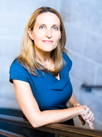 Woman with light brown hair wearing a blue top, leaning on a railing against a blurred gray background.