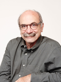Smiling older man with glasses and mustache wearing a dark button-up shirt against a plain background.