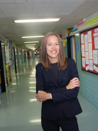 Smiling woman with shoulder-length brown hair stands in a school hallway with arms crossed.