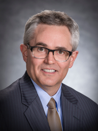 Middle-aged man with gray hair, glasses, wearing a suit, light blue shirt, and gold tie smiling at camera.