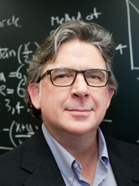 Middle-aged man with gray hair and glasses standing in front of a chalkboard with math equations.