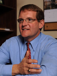 Man with glasses wearing a blue shirt and red tie, seated indoors gesturing with his hands.