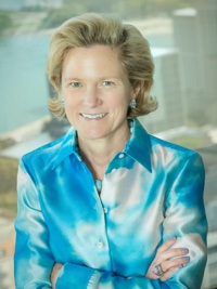 Smiling woman with short blond hair wearing blue patterned blouse standing indoors by a window.