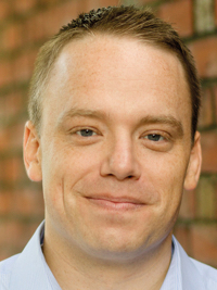 Close-up of a smiling man with short hair wearing a light blue collared shirt against a brick wall.