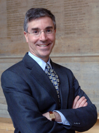 Man in suit and tie with glasses, smiling and standing with arms crossed against a beige wall.