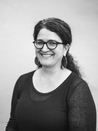 Smiling woman with glasses and long hair pulled back wearing a dark top against plain background.