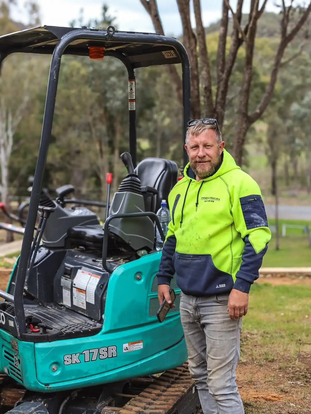 Man wearing a neon yellow and navy hoodie standing next to a teal SK 17SR excavator outdoors.