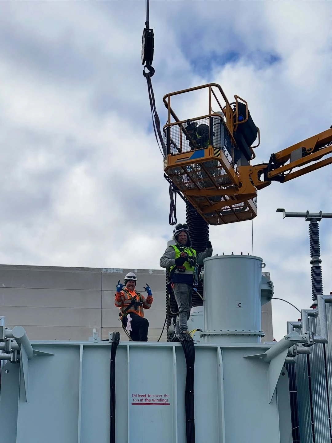 Two workers in safety gear on top of electrical equipment, one in a yellow lift and the other standing, with a cloudy sky background.