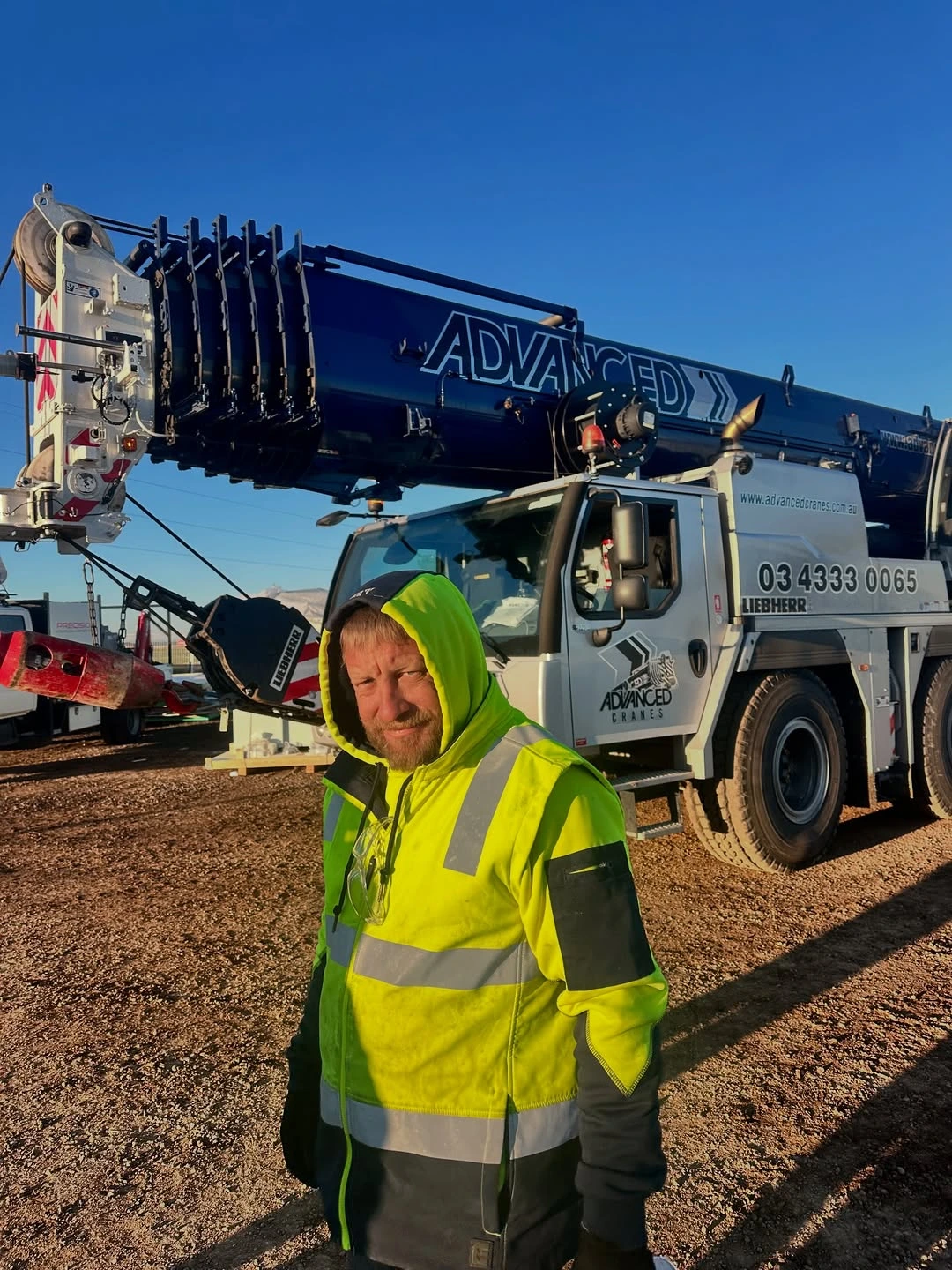 Man in a bright yellow and black high-visibility jacket with hood, standing in front of a large crane truck labeled 'Advanced Cranes' under a clear blue sky.