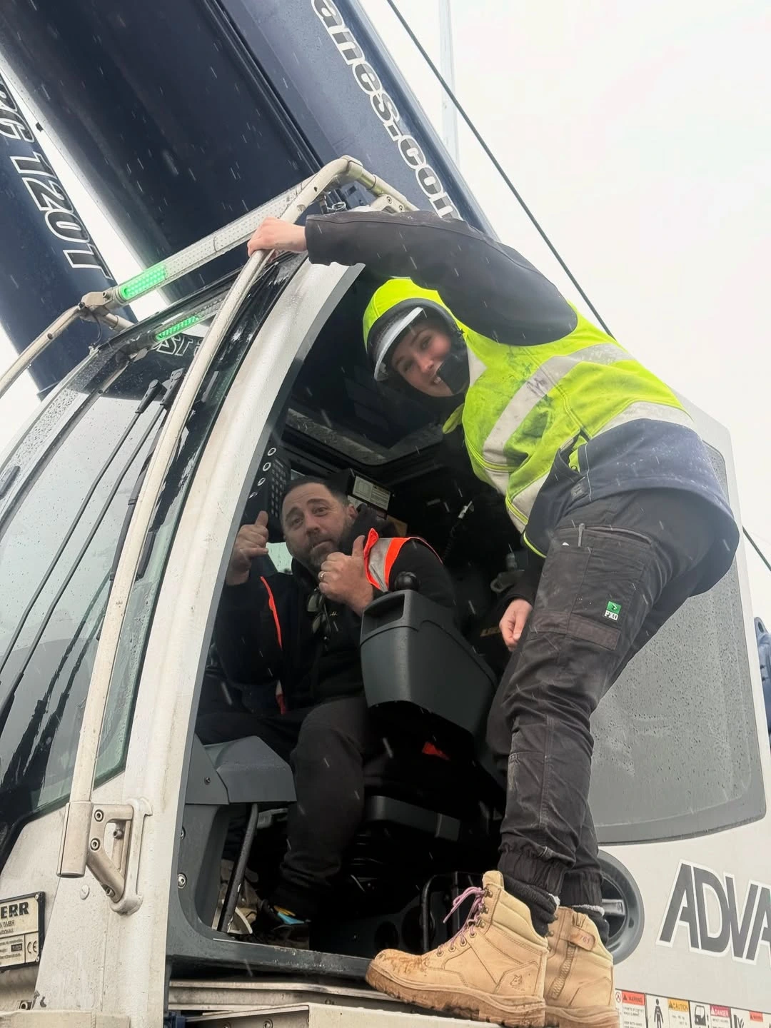 Two construction workers in safety vests inside and at the door of a heavy machinery cab on a rainy day.