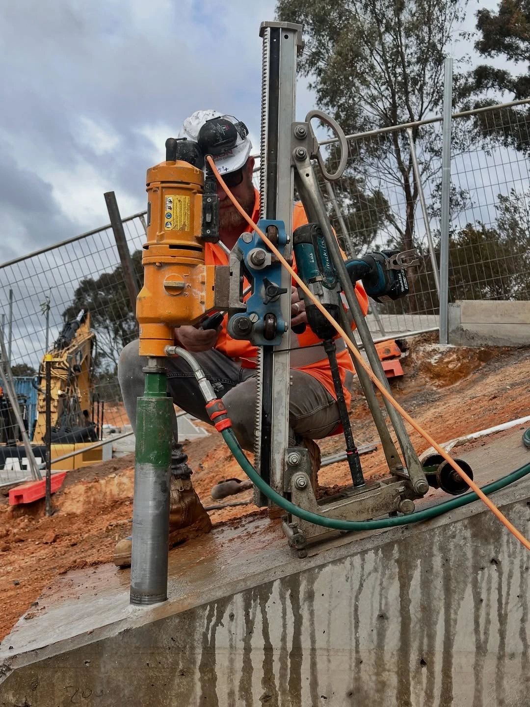 Construction worker in an orange vest operating a core drilling machine on a concrete surface at a construction site.