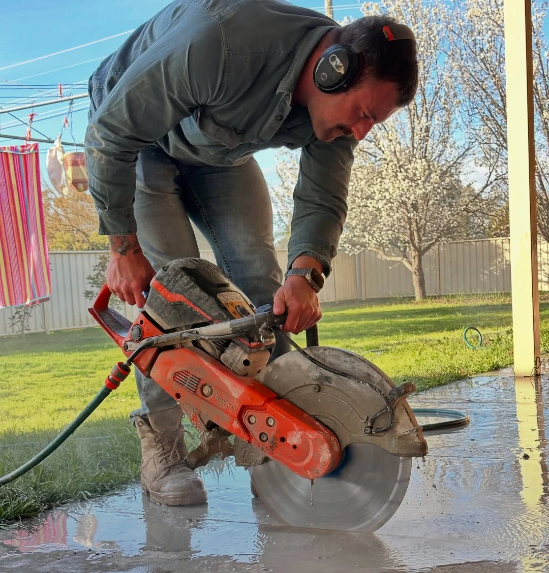 Man wearing headphones using an orange electric saw to cut wet concrete on a patio outdoors.