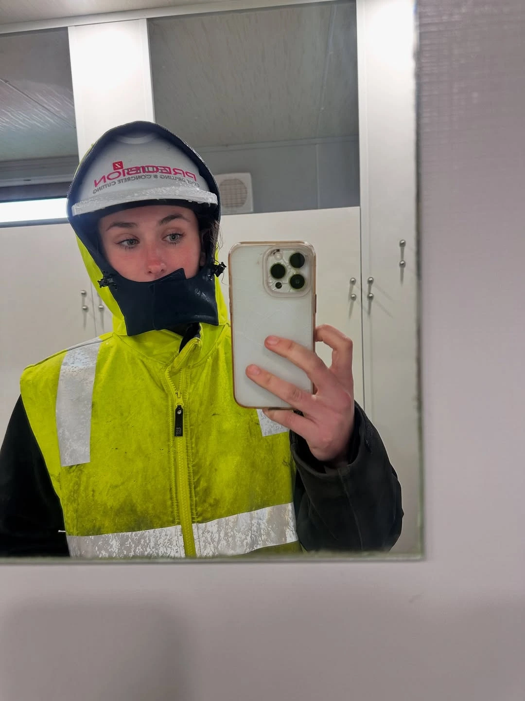 Person wearing a white helmet with a visor and a yellow high-visibility jacket taking a mirror selfie with a smartphone.