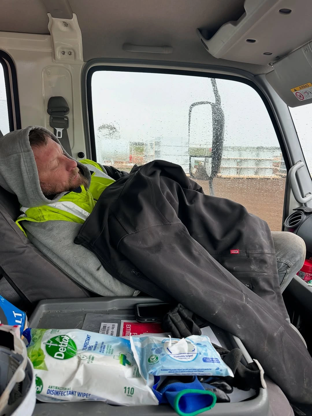 Man in a gray hoodie and yellow safety vest sleeping inside a vehicle, covered with a black jacket on the passenger seat.