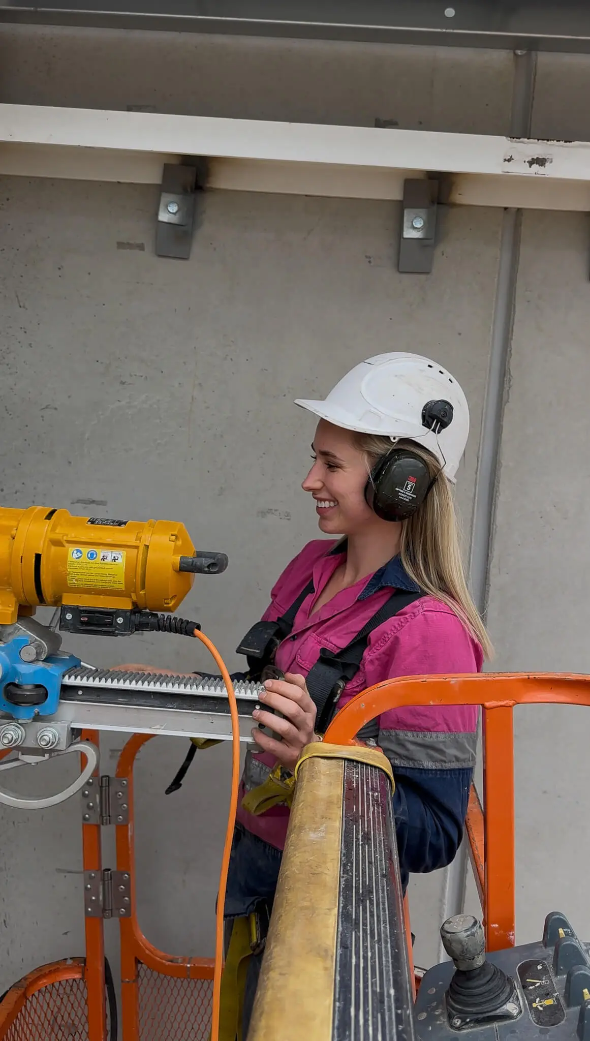 Smiling female construction worker wearing a white hard hat and ear protection operating yellow machinery on an orange lift platform.
