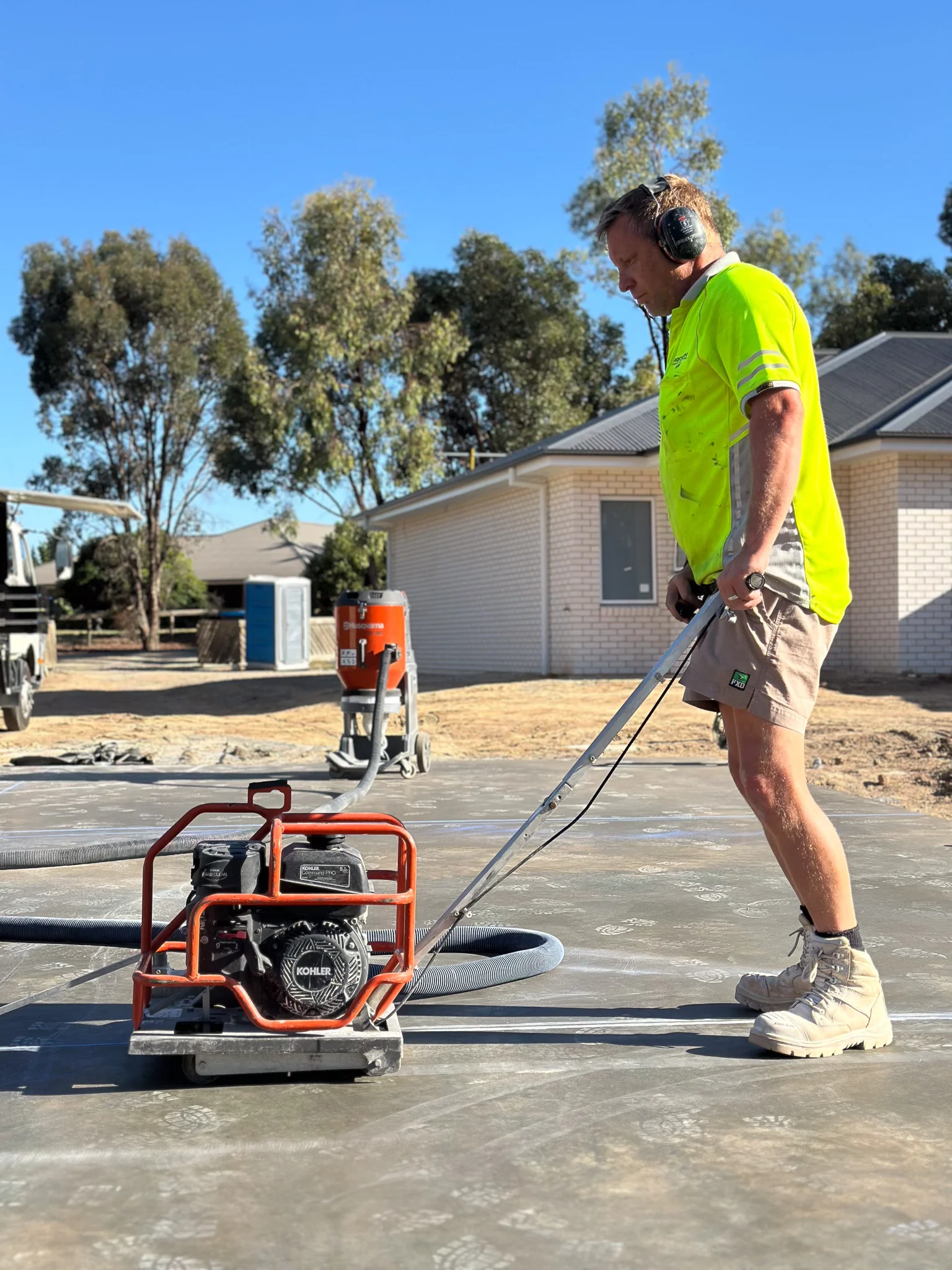 Construction worker in a neon yellow shirt and safety boots operating a concrete power trowel on a freshly poured concrete slab.