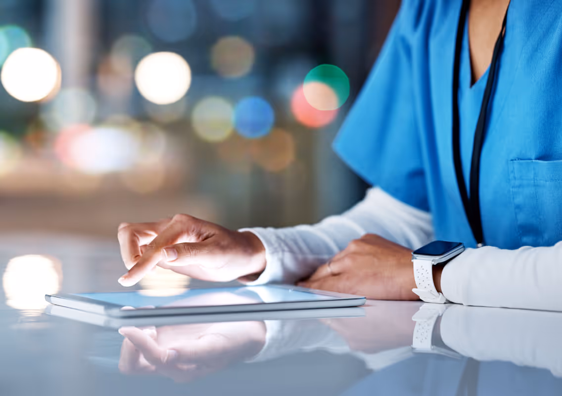 Healthcare professional in blue scrubs using a tablet on a reflective surface.