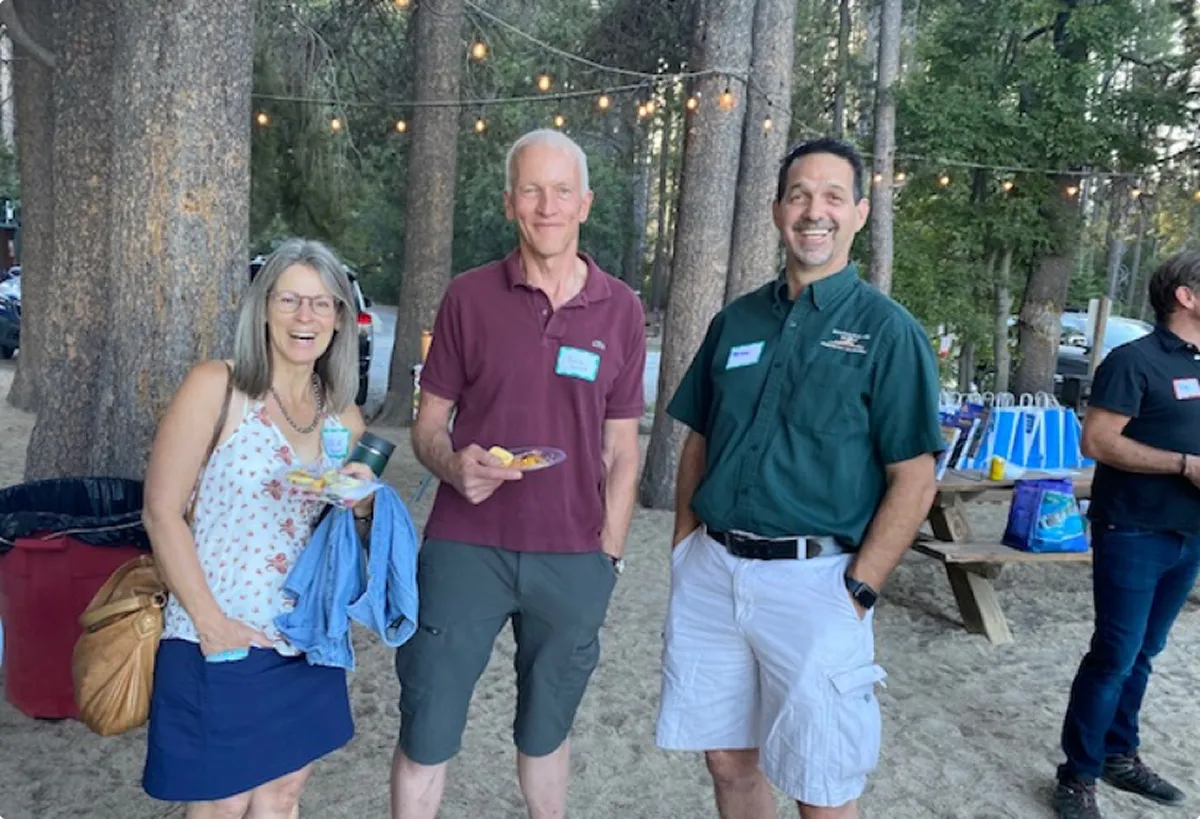 Three people smiling outdoors at a picnic area with string lights and trees in the background, holding plates of food.