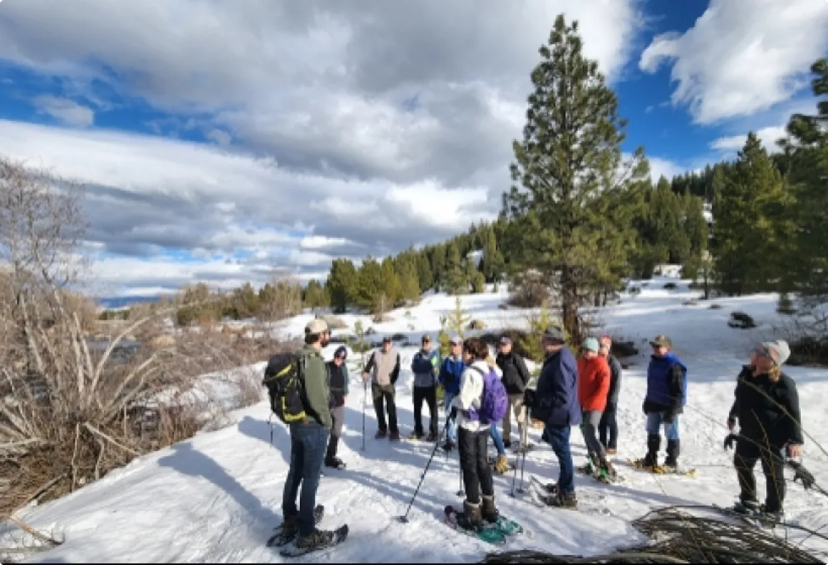 Group of people wearing snowshoes and winter clothing standing on snowy terrain with trees and a partly cloudy sky.