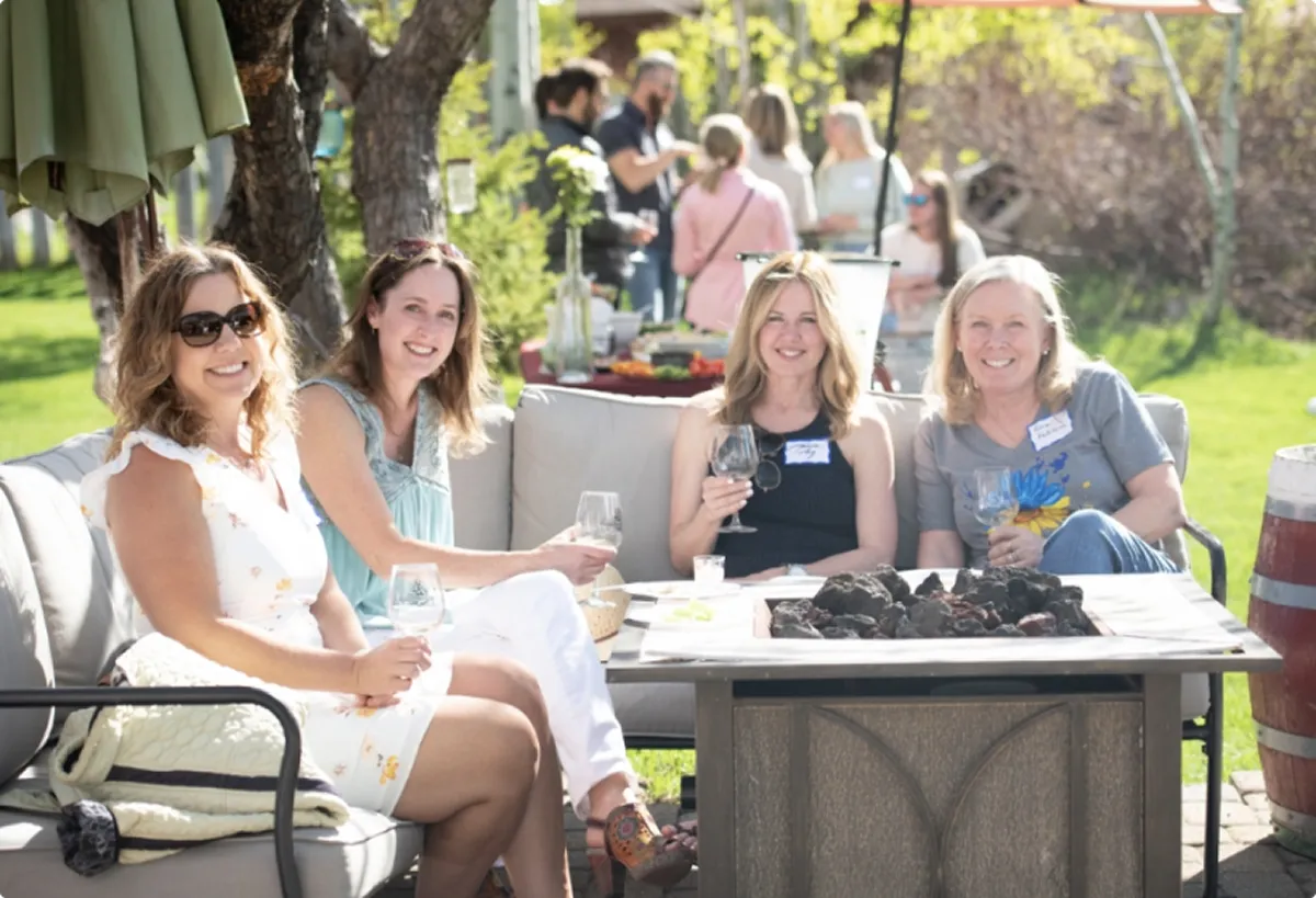 Four women sitting outdoors on a couch around a fire pit, holding wine glasses and smiling.