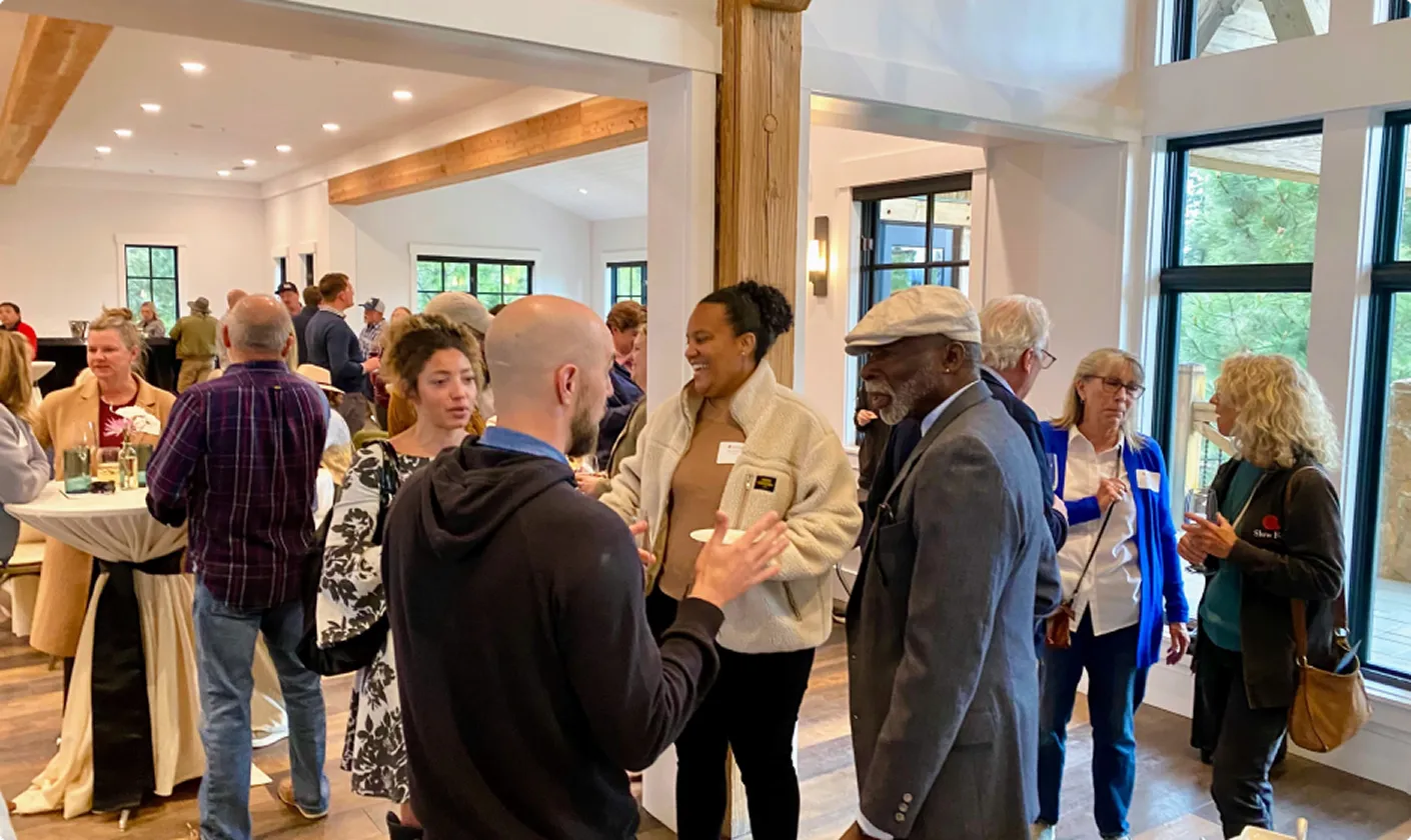 Group of diverse people engaged in conversation inside a bright room with large windows and wooden beams.