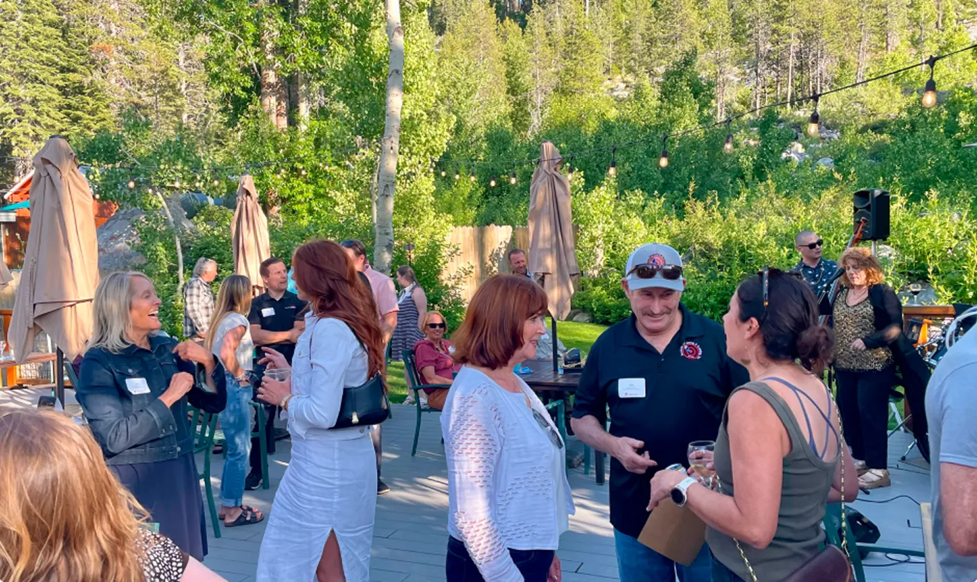 Group of people socializing outdoors on a sunny day with trees and string lights in the background.