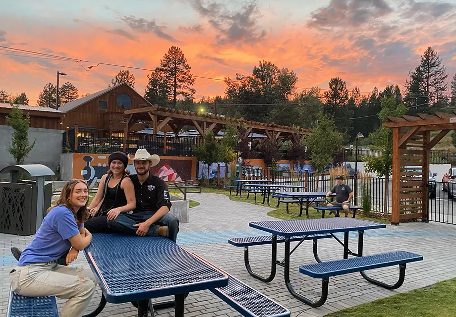 Three people sitting on and beside a blue metal picnic table in a park at sunset, with colorful sky and trees in the background.