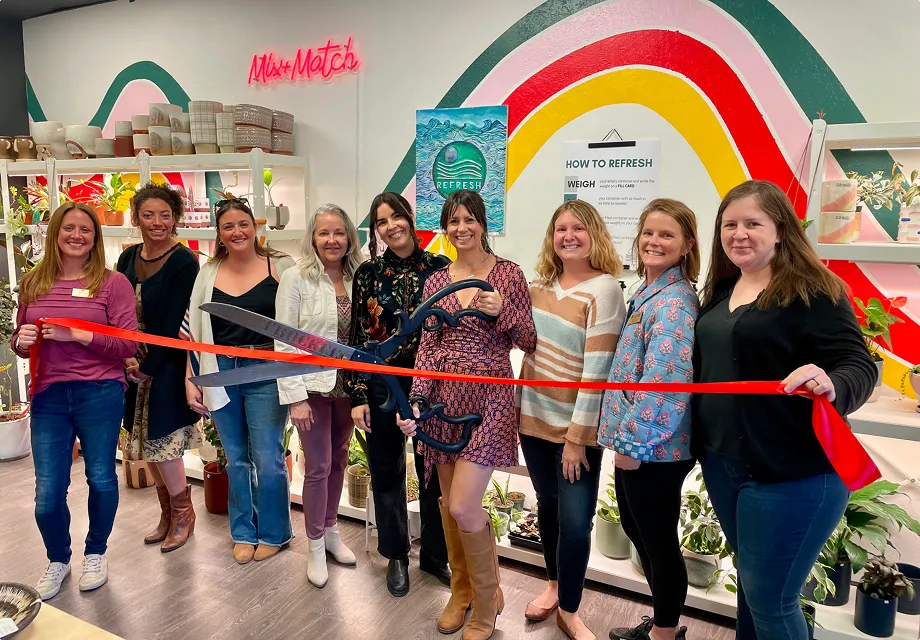 Group of nine smiling women standing indoors holding a large pair of scissors cutting a red ribbon during a ribbon-cutting ceremony.