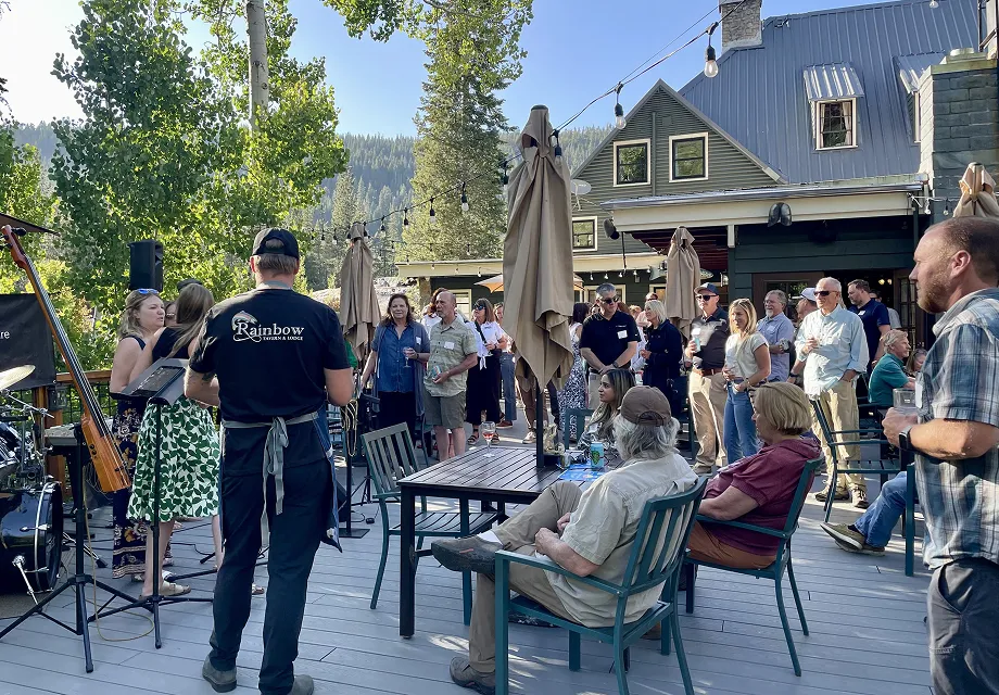 Outdoor gathering with people socializing on a deck, some seated and others standing near tables, with a live band setup and a building with umbrellas and string lights in the background.