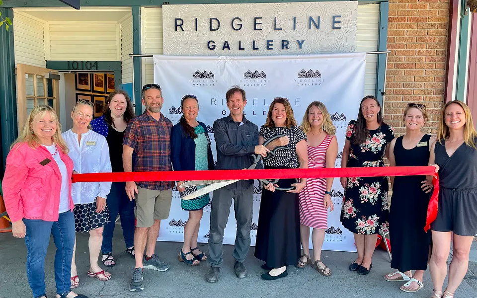 Group of people standing in front of Ridgeline Gallery holding a large pair of scissors to cut a red ribbon during a ribbon-cutting event.