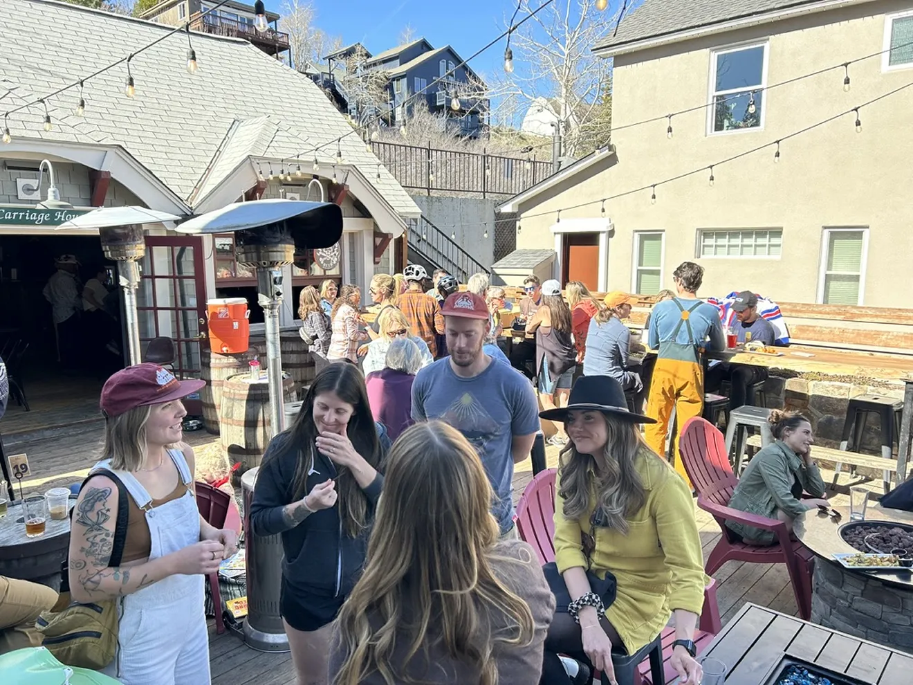 Outdoor gathering with people socializing on a sunny patio featuring wooden tables, string lights, and a fire pit.