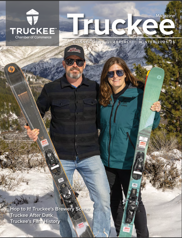 Man and woman standing on snow-covered ground holding skis with snow-capped mountains and pine trees in the background.
