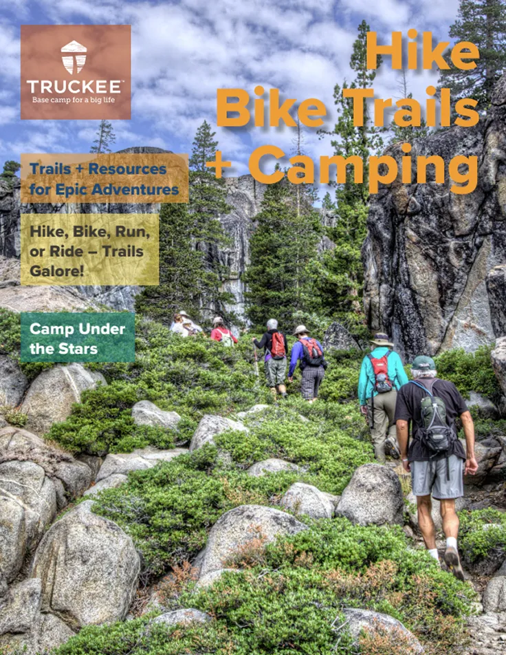 Group of hikers walking up a rocky trail surrounded by green shrubs and tall pine trees under a partly cloudy sky.