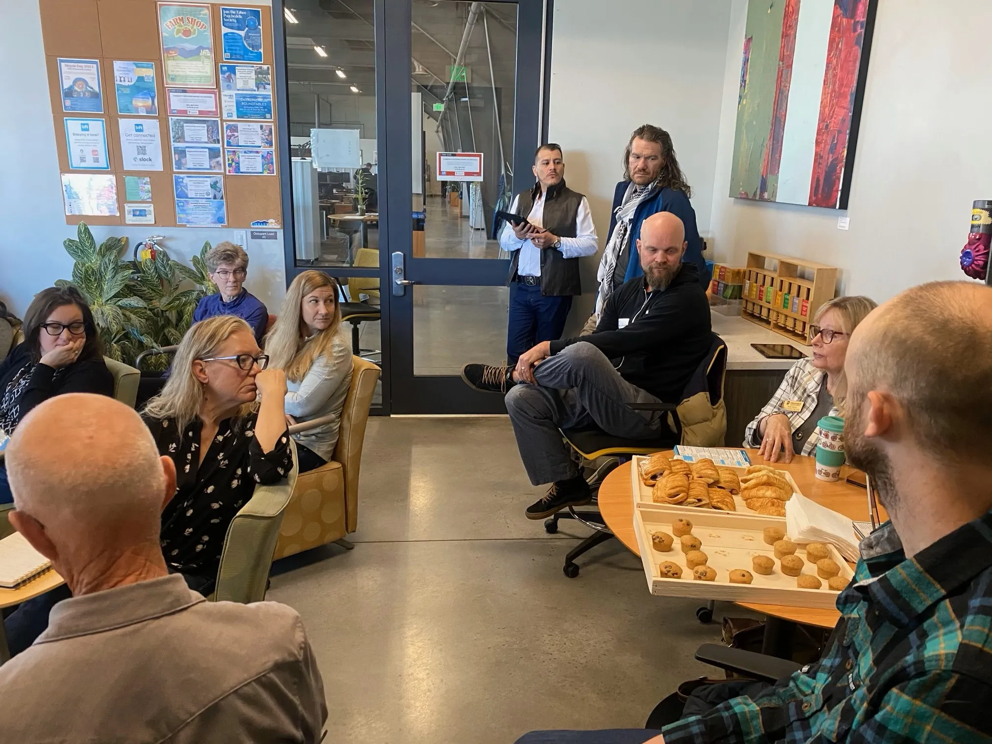 Group of people seated and standing in a casual meeting room with trays of pastries on a table.