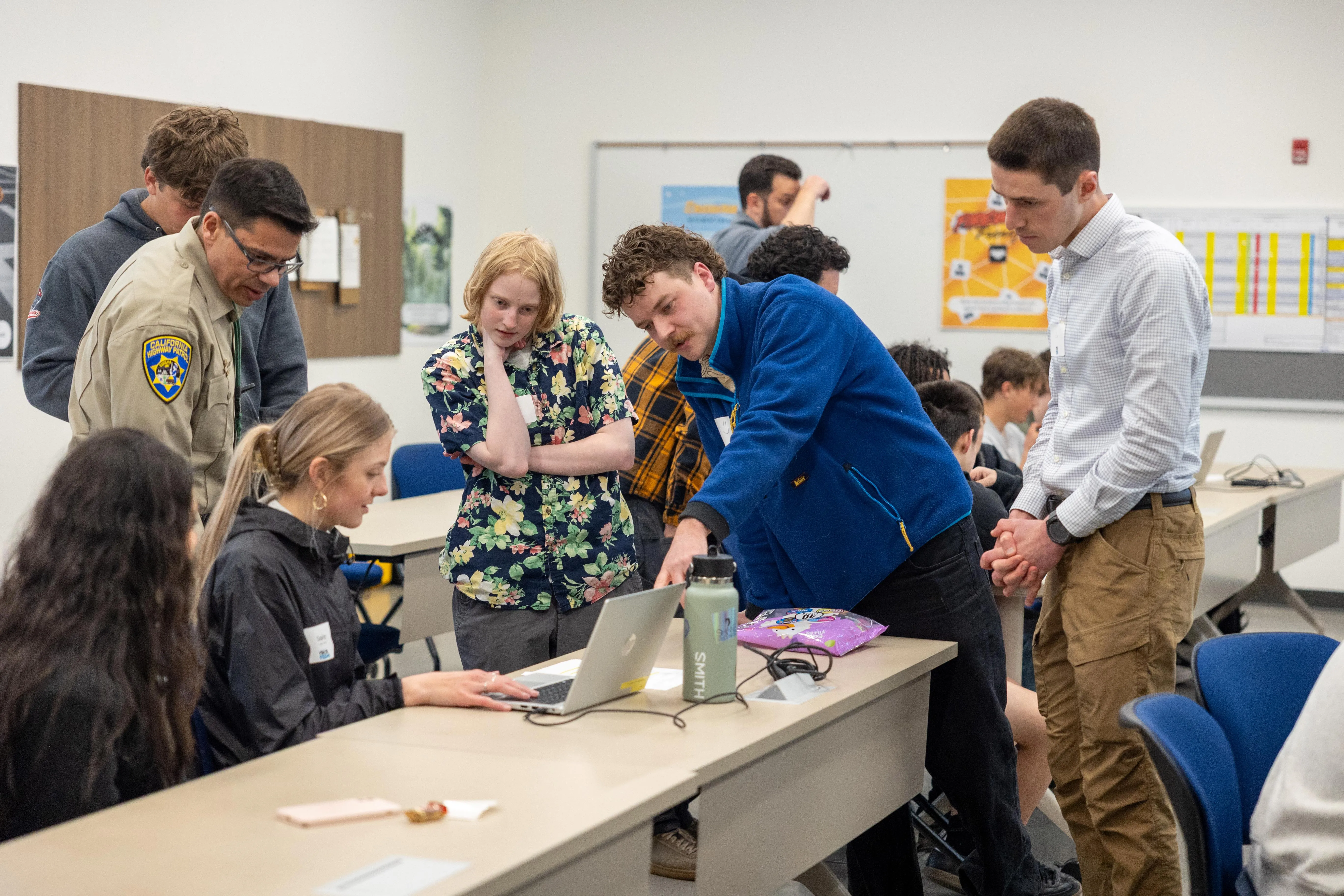 Group of diverse people, including a California Highway Patrol officer, gathered around a laptop in a classroom setting, discussing and focused on the screen.