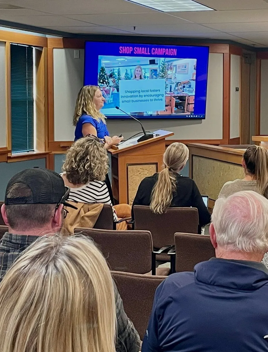 Woman in blue speaking at a podium to an audience in a conference room, with a screen behind her showing a Shop Small Campaign message about supporting local small businesses.