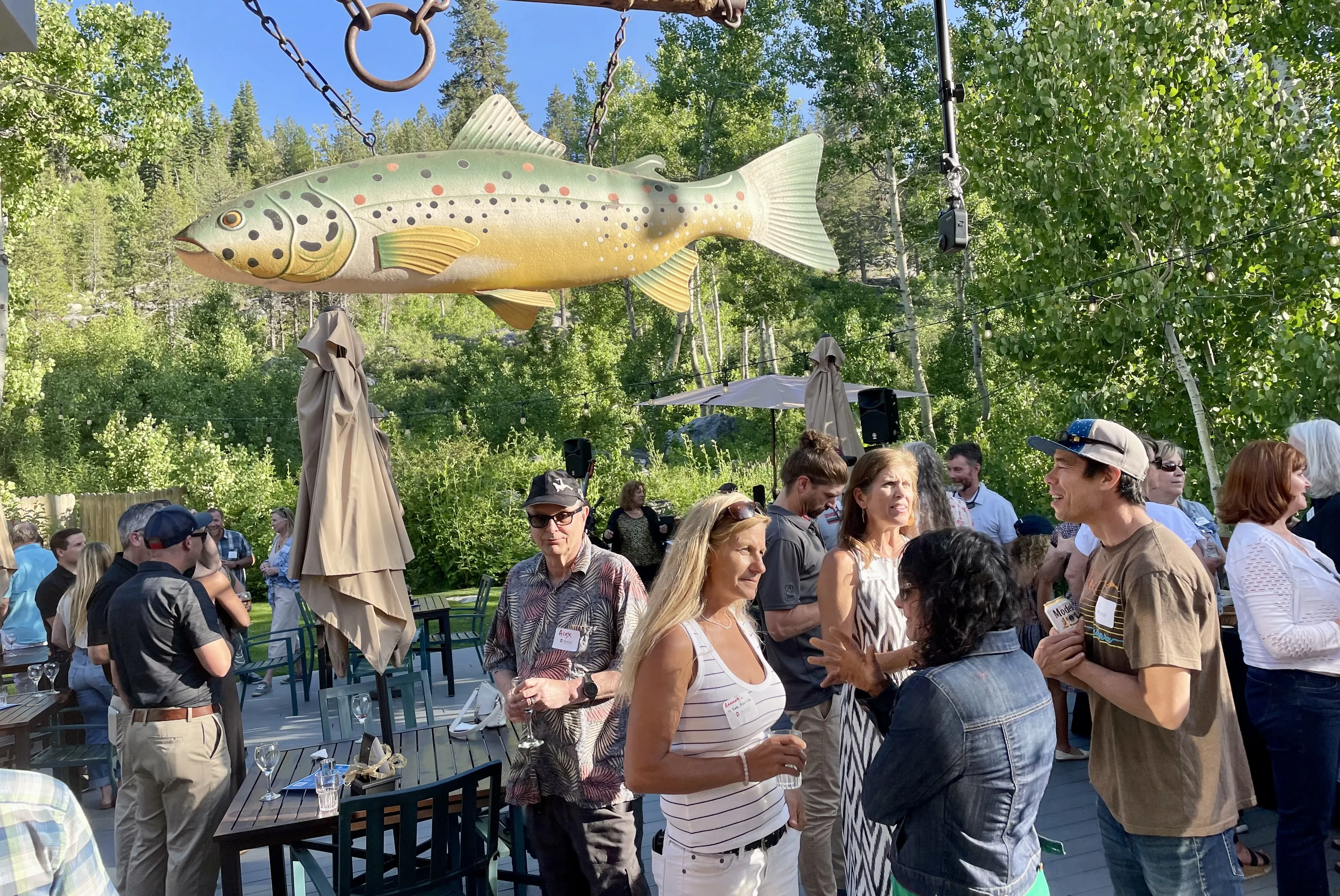 Outdoor gathering of people socializing under a large hanging fish decoration with trees and clear blue sky in the background.