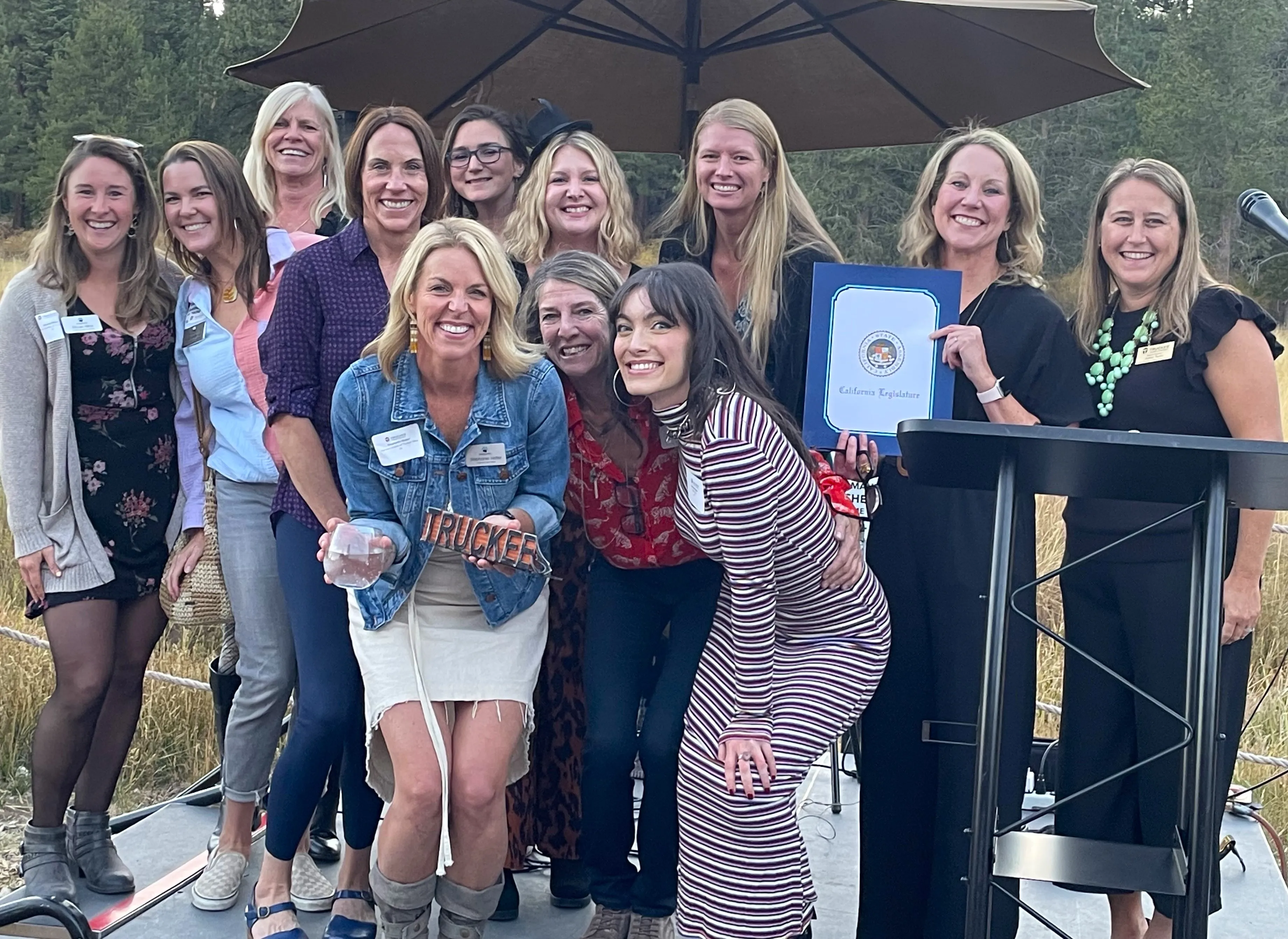 Group of smiling women posing outdoors on a small stage under a large umbrella, one holding a California Legislature certificate and another holding a glass award labeled 'Truckee'.