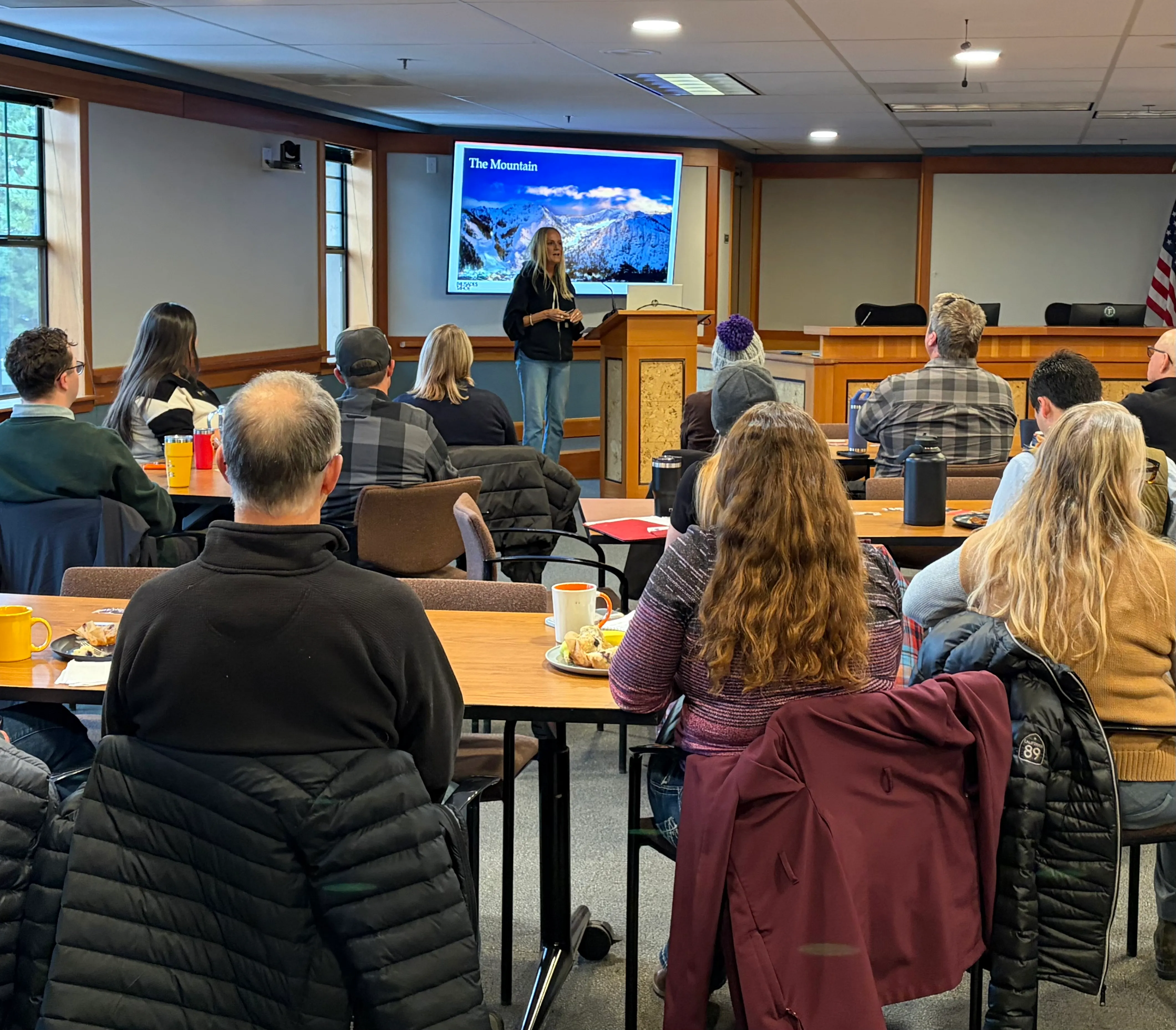 A woman giving a presentation titled 'The Mountain' to an audience seated at tables in a conference room.