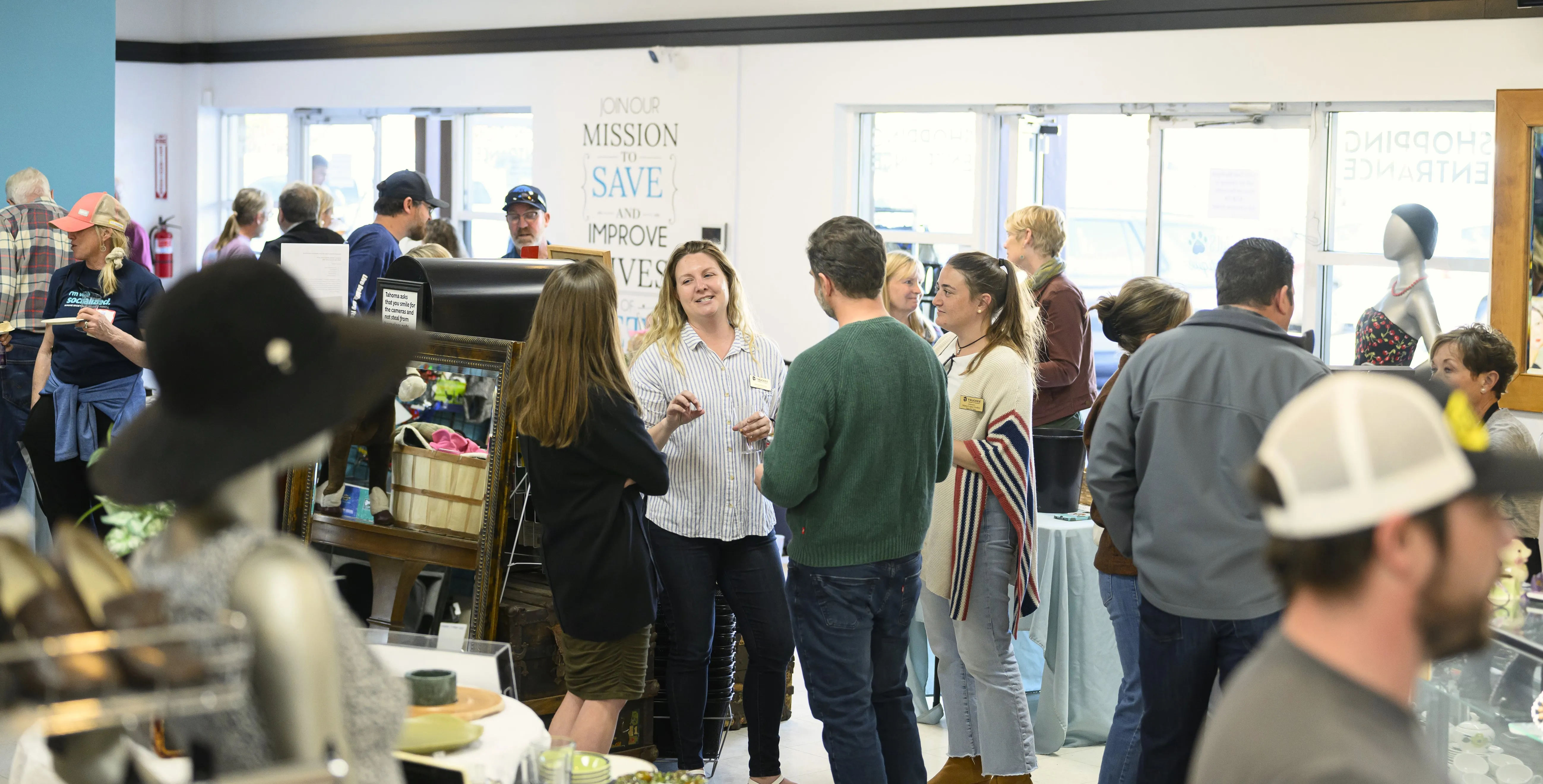 People socializing inside a bright store with clothing racks, a mannequin, and a sign on the wall about a mission to save and improve lives.