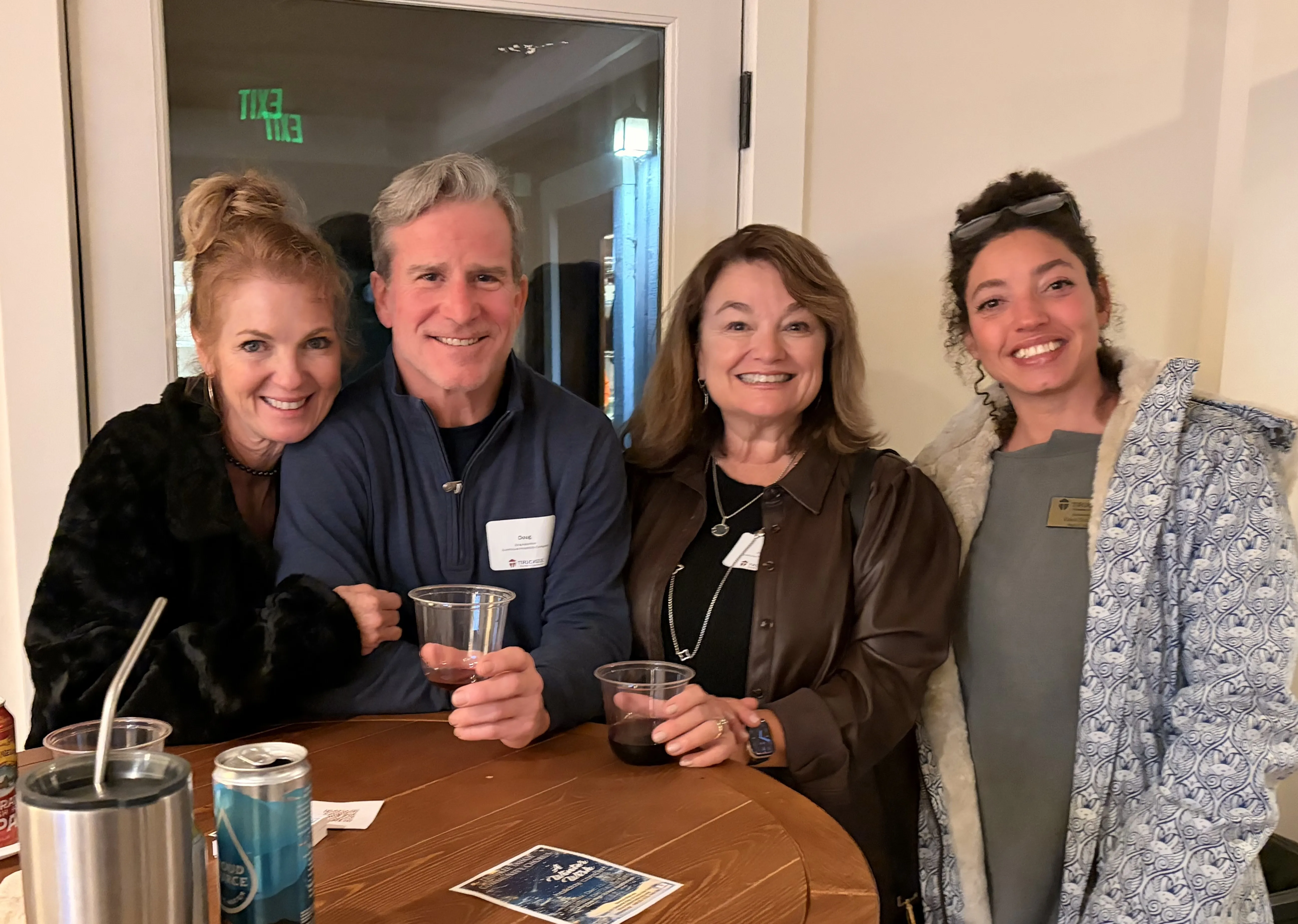 Four people smiling indoors around a wooden table with drinks, two women on each side and a man in the middle.