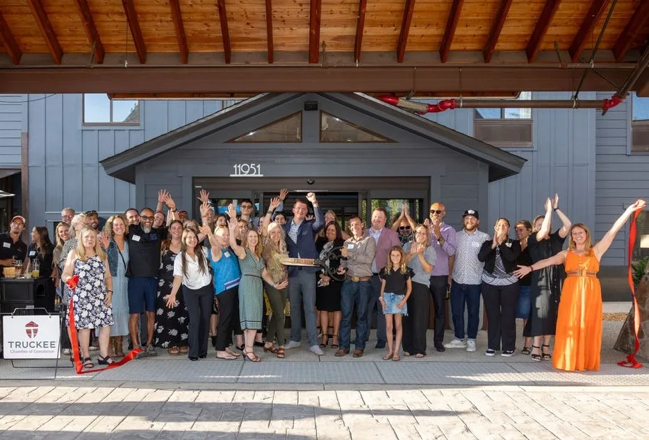 Group of people gathered outside a building with sign '11951' celebrating a ribbon-cutting event, with two women holding red ribbon ends and a man holding scissors in the center.