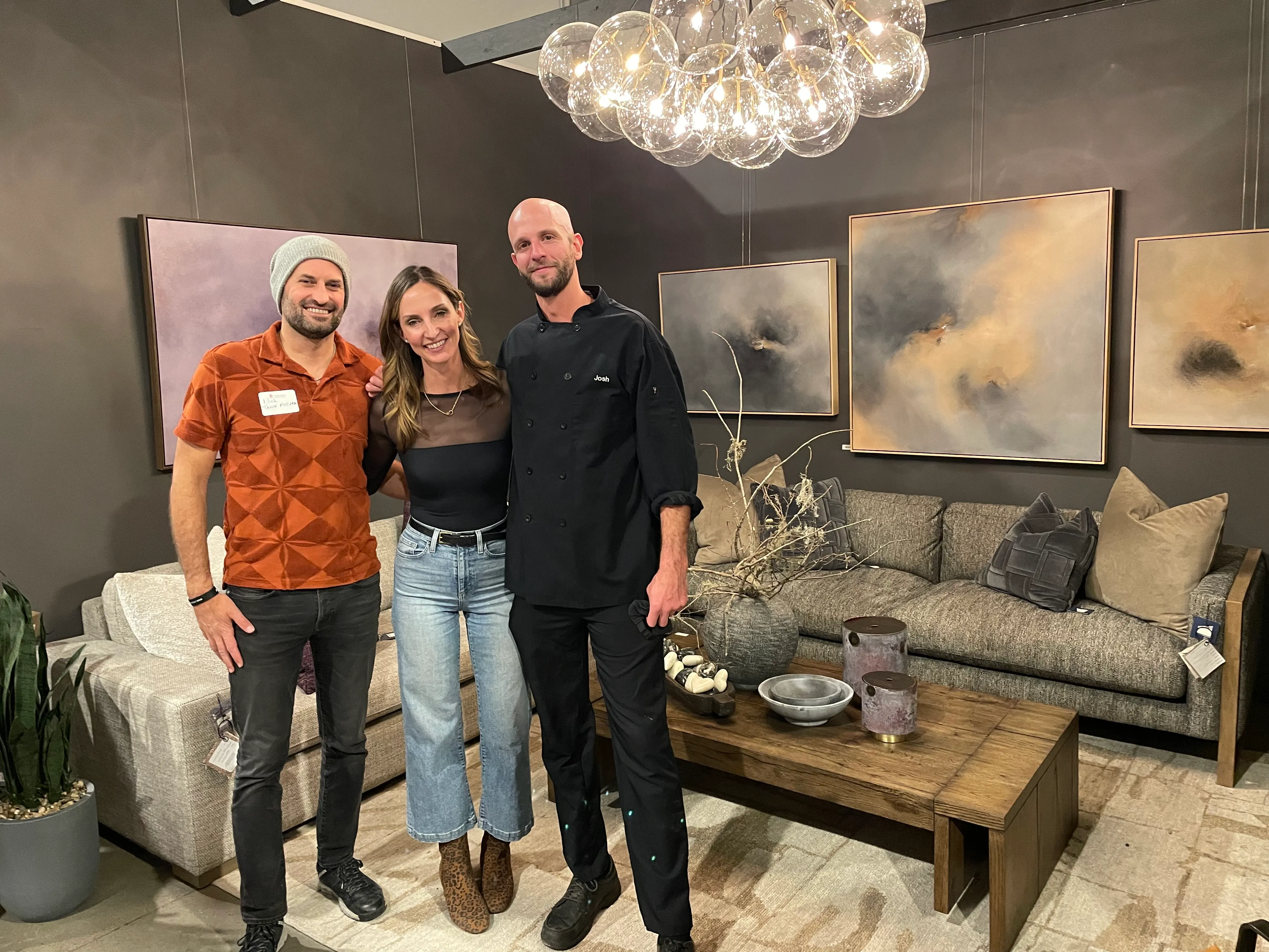 Three people standing closely in a modern living room with grey walls, abstract paintings, a wooden coffee table, and a stylish chandelier overhead.