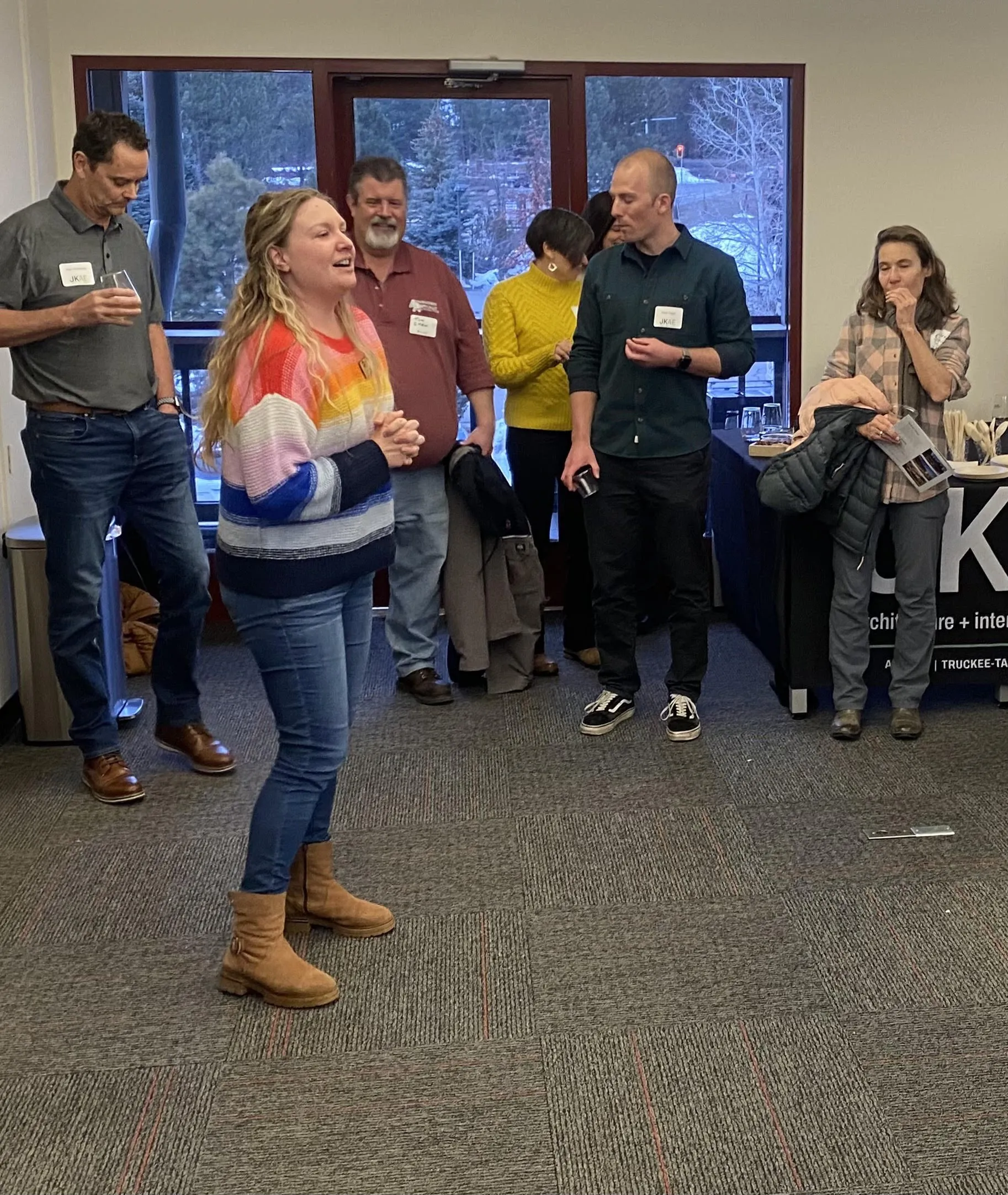 A woman in a colorful striped sweater speaks to a group of five people standing indoors near large windows.