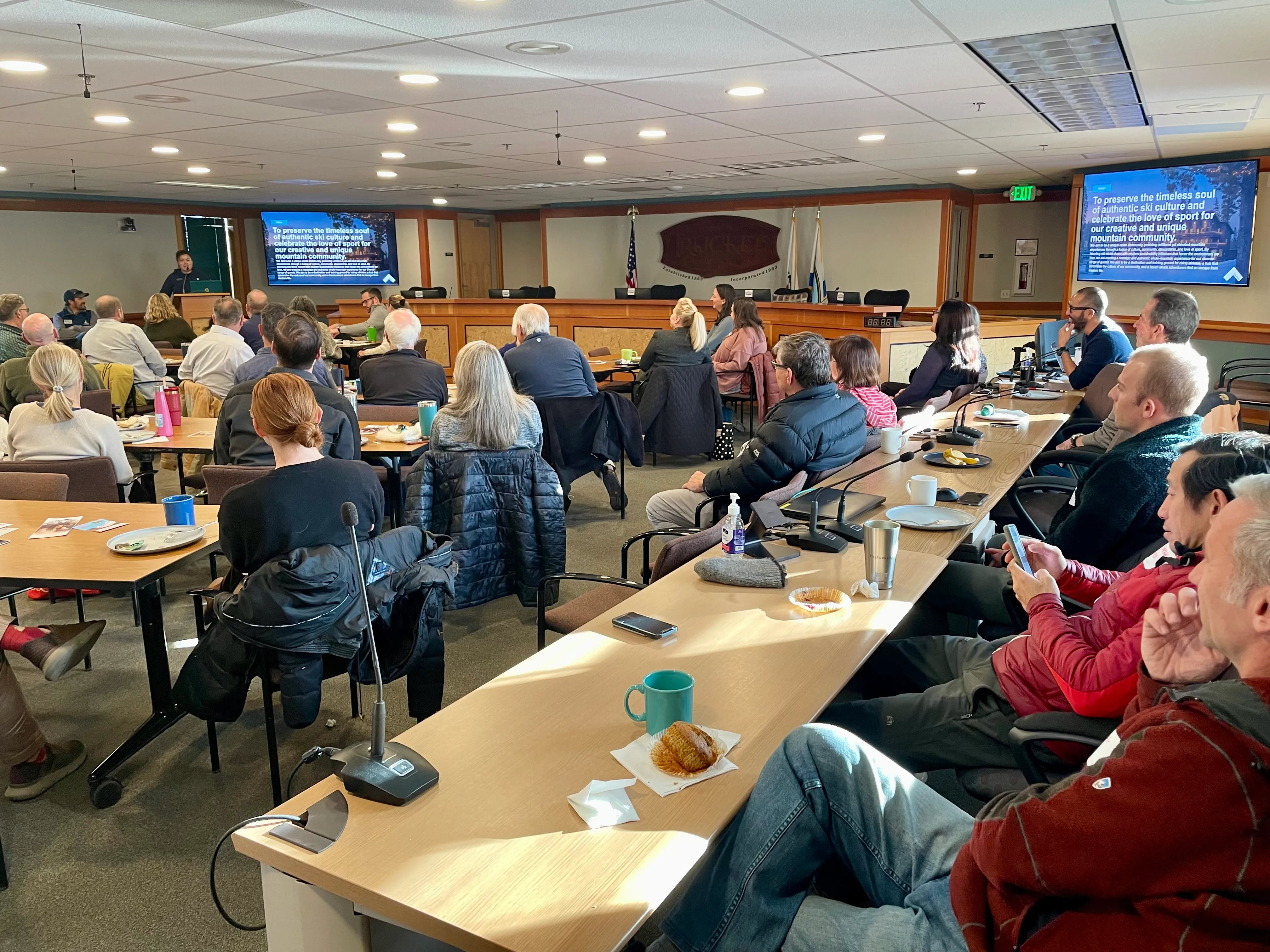 People seated in a conference room attentively listening to a speaker presenting with slides on two large screens.