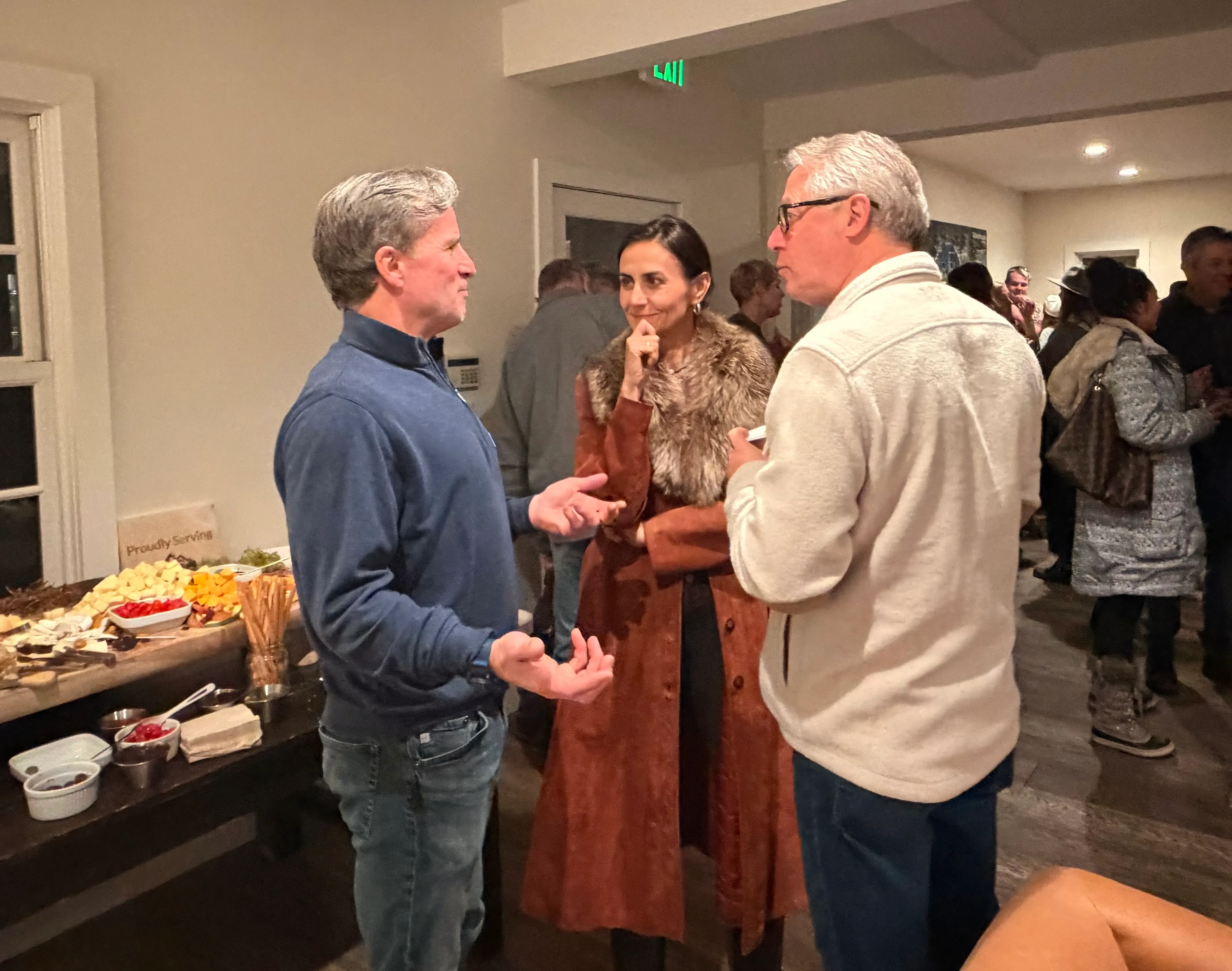 Three adults engaged in conversation at an indoor gathering with a table of cheeses and snacks nearby.