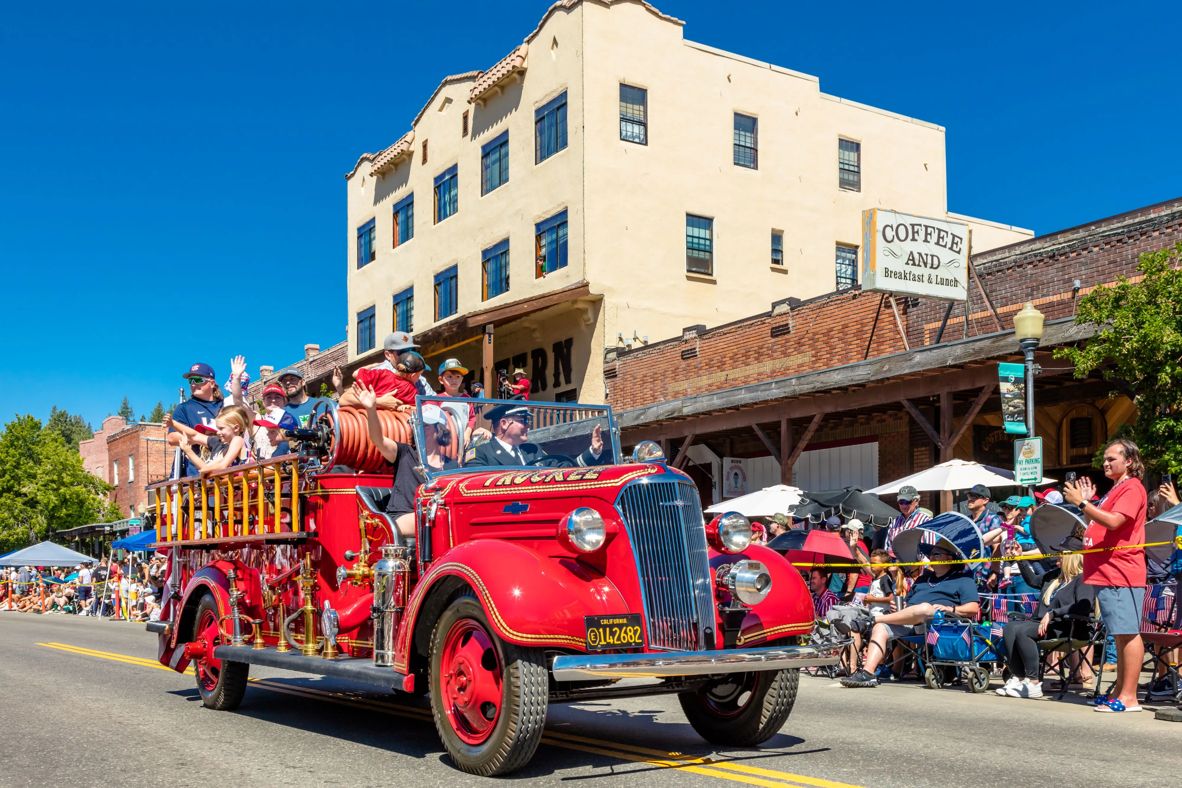 Vintage red fire truck carrying people waving in a sunny parade with spectators seated and standing along the street.